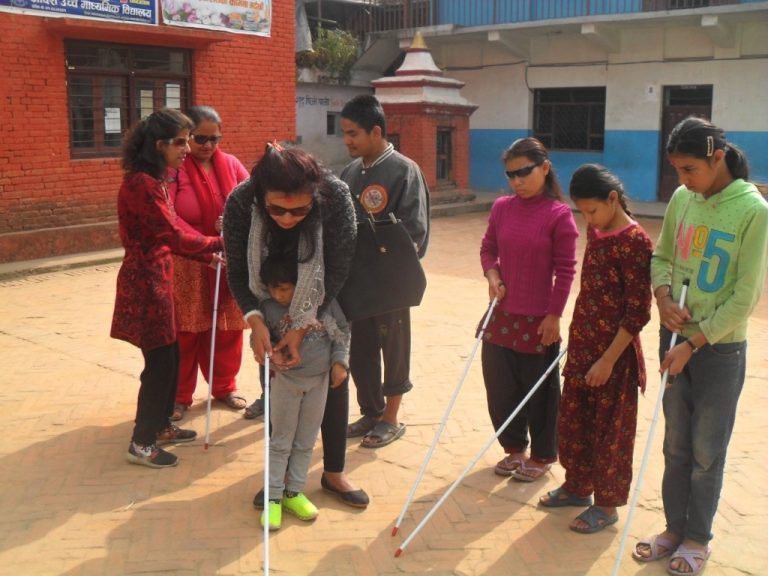 A trainer is teaching a visually impaired child how to use the white cane. Other participants are also seen in the photo.