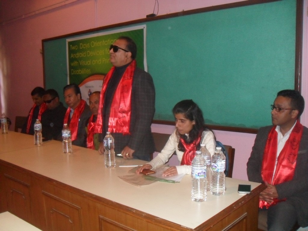 A person is standing and speaking while others are sitting beside him