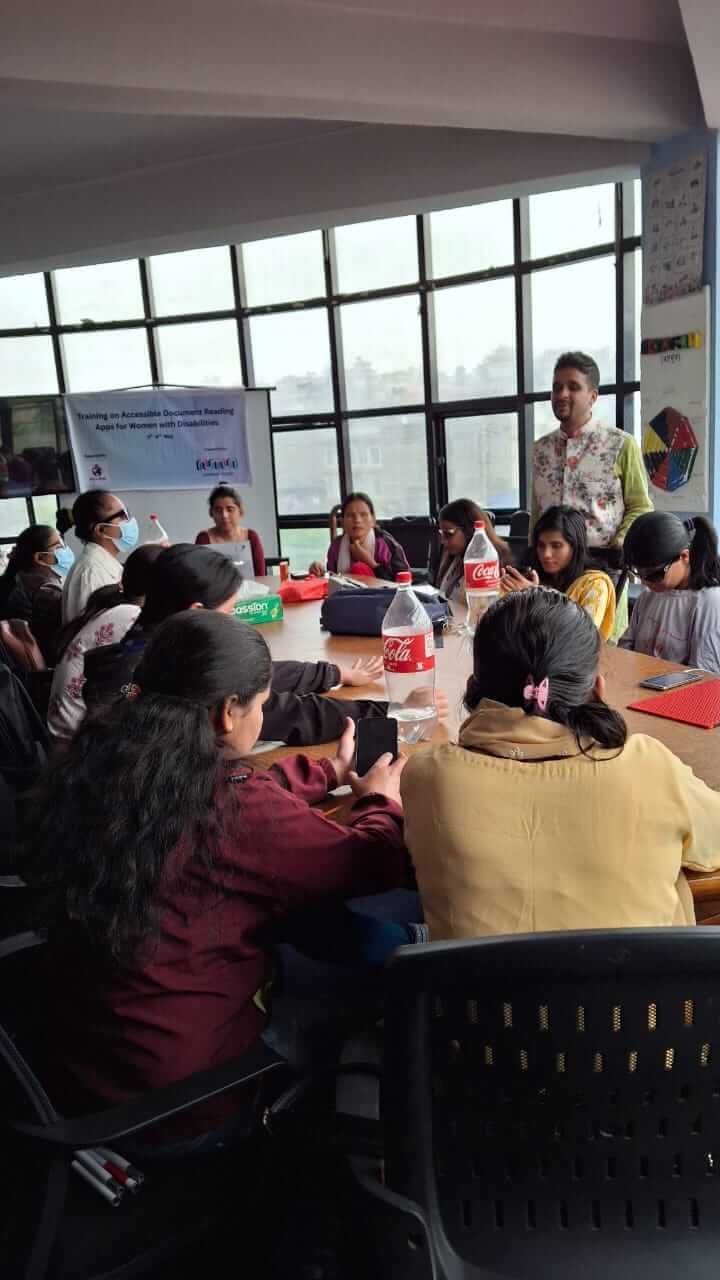 A group of women sit around a conference table, attentively using smartphones during a training session. A trainner stands at the front, speaking to the participants. A banner in the background reads “Training on Accessible Document Reading Apps for Women wi