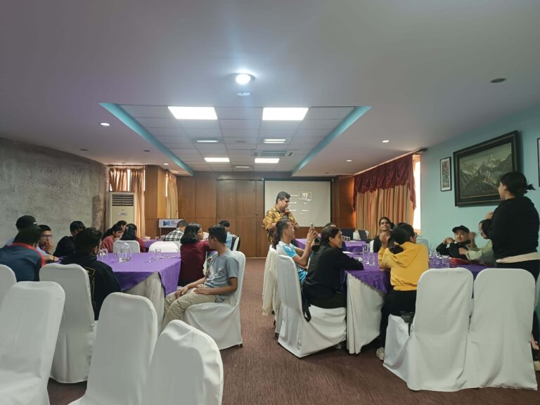 A conference room filled with young people seated at tables with purple tablecloths, some interacting and one person standing at the front speaking into a microphone.