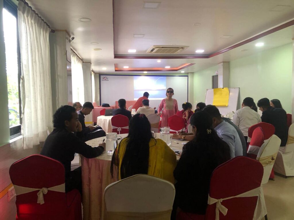 A group of people is seated at round tables in a conference room, with Laxmi Nepal standing in the center, wearing a pink shirt and dark sunglasses. She is gesturing with her hands while speaking to the group. A projector screen with a presentation is visible in the background.