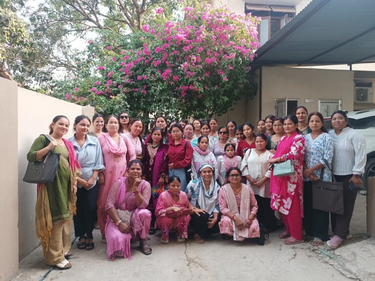 A group of women, dressed in a variety of clothing, are posing together for a group photo outdoors. They are standing and crouching in front of a building, with a vibrant pink bougainvillea plant visible behind them.