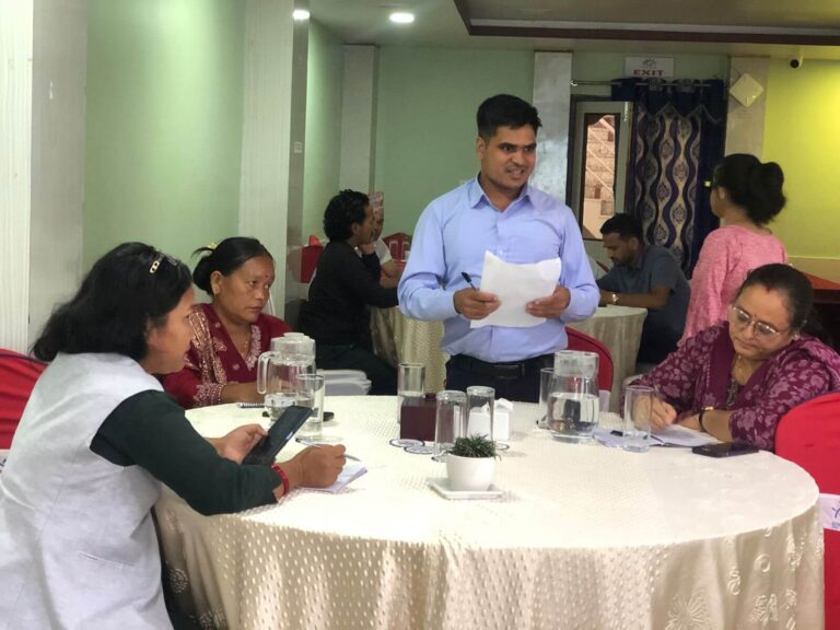 A man in a light blue shirt, stands and speaks to a group of four women seated around a circular table during a session, holding papers in his hand.