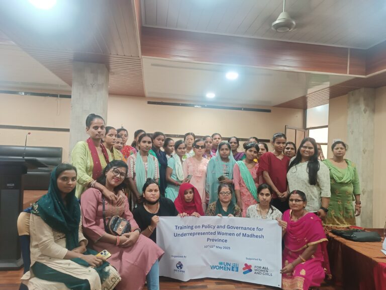 A group of women stand and sit together, posing for a photo indoors. Several women in the front row are holding a white banner that reads "Training on Policy and Governance for Underrepresented Women of Madhesh Province," with the dates 10th-11th May 2025.