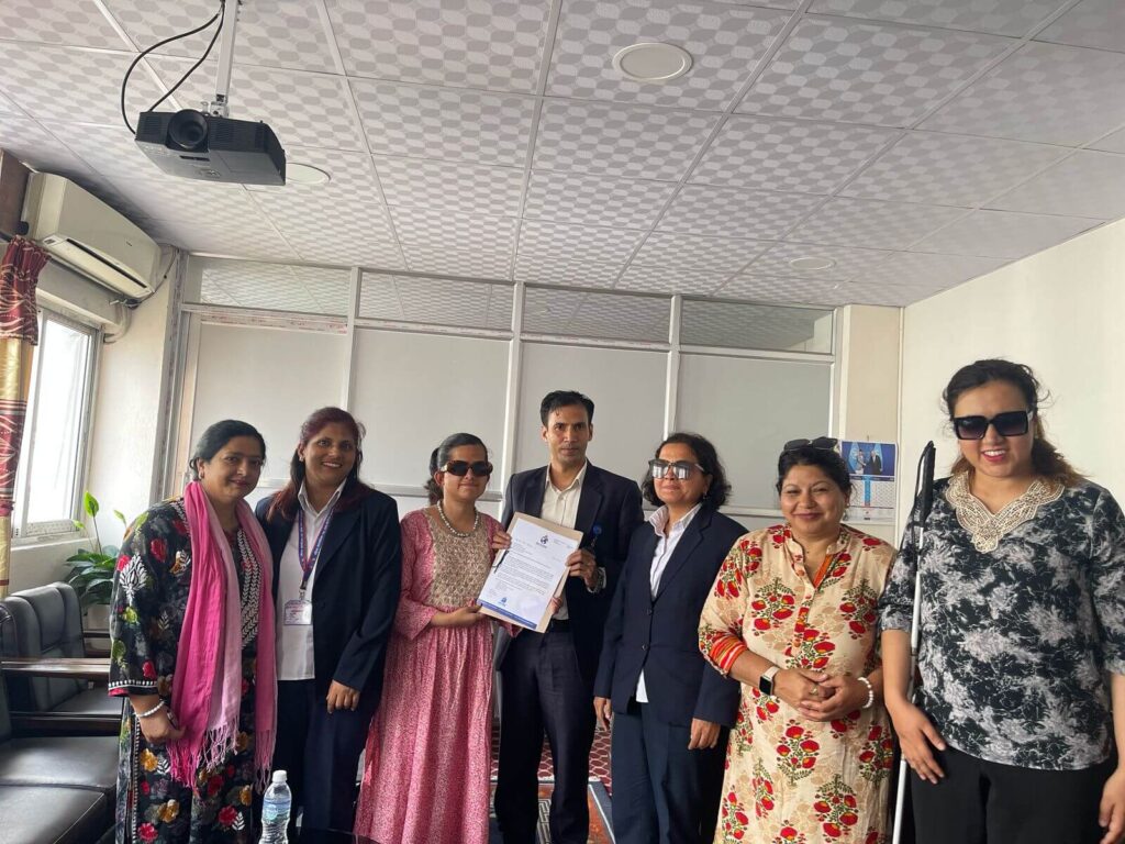 General Secretary Gyanmani Ghimire is in the middle, holding a proposal. He is with Ms. Kamala Ghimire, Head of the Disability Rights Promotion Division, and Ms. Neera Adhikari, Head of the Social Coordination Division, along with representatives of organizations for women with disabilities. The representatives are submitting the proposal, and one of them, Laxmi Nepal, is standing next to Mr. Ghimire. One of the women on the far right is holding a white cane.