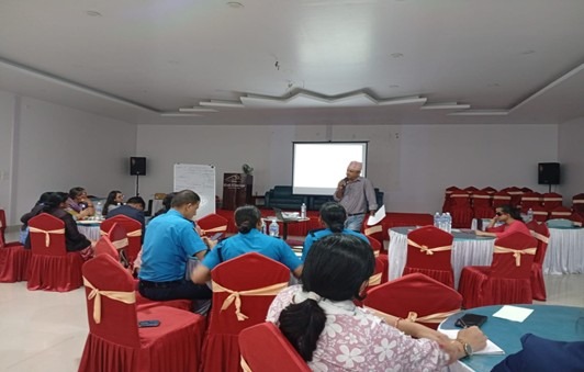 A conference room scene where a man in a light shirt and traditional Nepali topi stands near a large, blank projector screen, addressing a seated audience. The attendees are seated at round tables covered in red cloths, and a few are wearing police uniforms. The room has high ceilings, speakers on either side of the screen, and a whiteboard to the left.