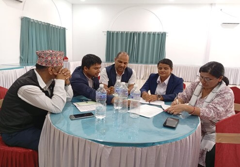 A group of five people, three men and two woman, sit around a round table covered with a light blue cloth. They appear to be engaged in a discussion, with papers and water bottles on the table. One man wears a traditional Nepali topi, and the woman on the right is writing. The room is a conference or banquet hall with light-colored curtains in the background.