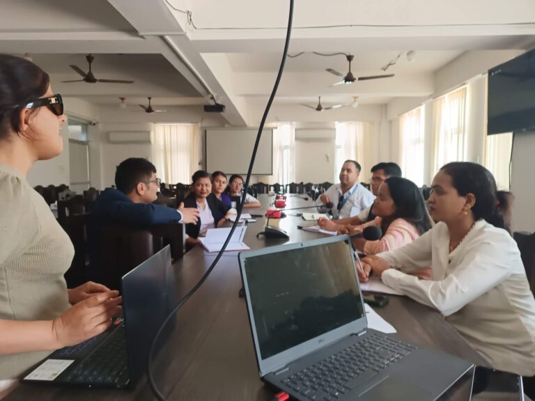 A group of people, including both men and women, are seated around a conference table in a meeting. Laxmi Nepal in the foreground is speaking while others listen and take notes. Laptops and notebooks are visible on the table.